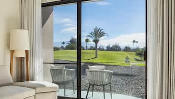 Una habitación con sofá, dos sillones de mimbre, una lámpara y un amplio ventanal corredera que da a un jardín ajardinado con una palmera y montañas al fondo.