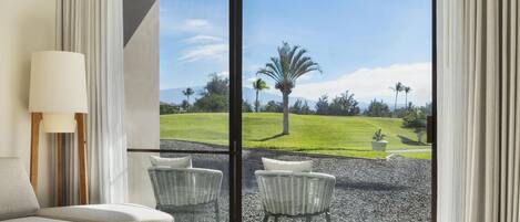 Una habitación con sofá, dos sillones de mimbre, una lámpara y un amplio ventanal corredera que da a un jardín ajardinado con una palmera y montañas al fondo.