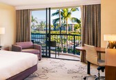 A hotel room with a bed, a desk, a chair, and a view of palm trees and a pool.