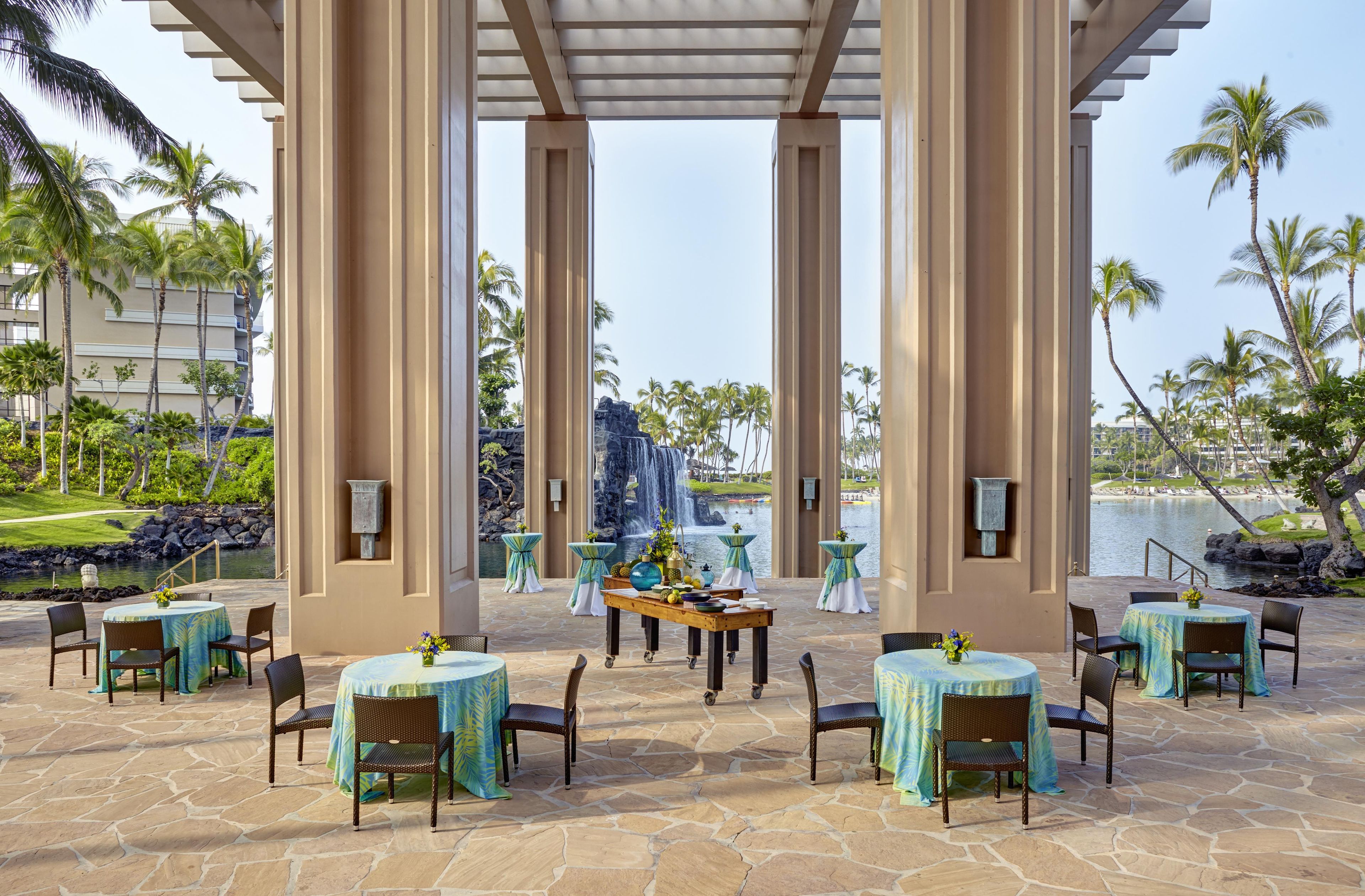 An outdoor seating area with tables and chairs, a waterfall view, and palm trees.