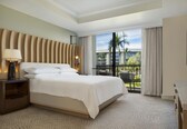 A hotel room with a large bed, a nightstand, a lamp, a desk, and a view of a building and palm trees through the window.
