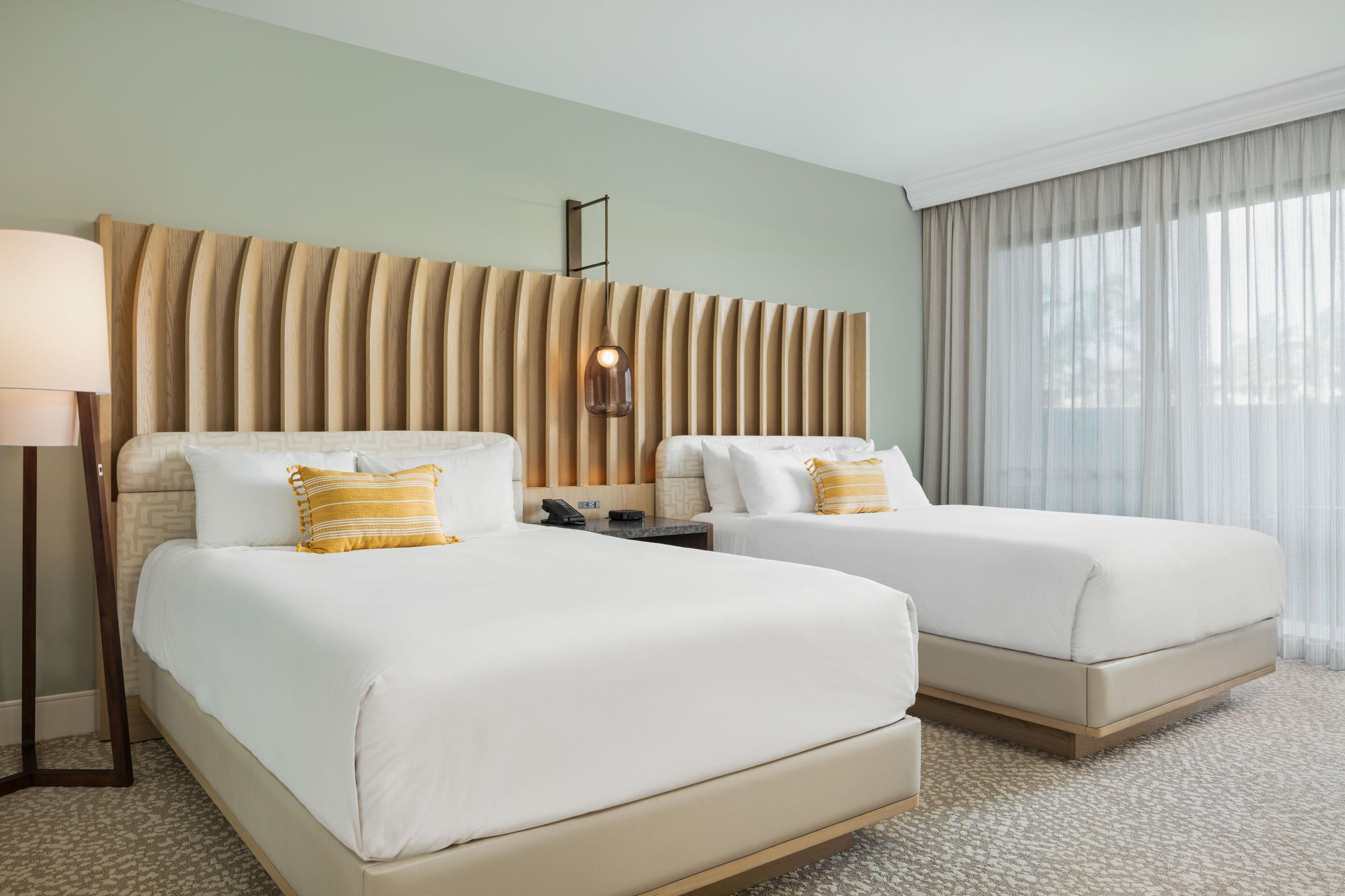 A hotel room with two beds, a wooden headboard, a bedside table with a lamp, and a large window with curtains.