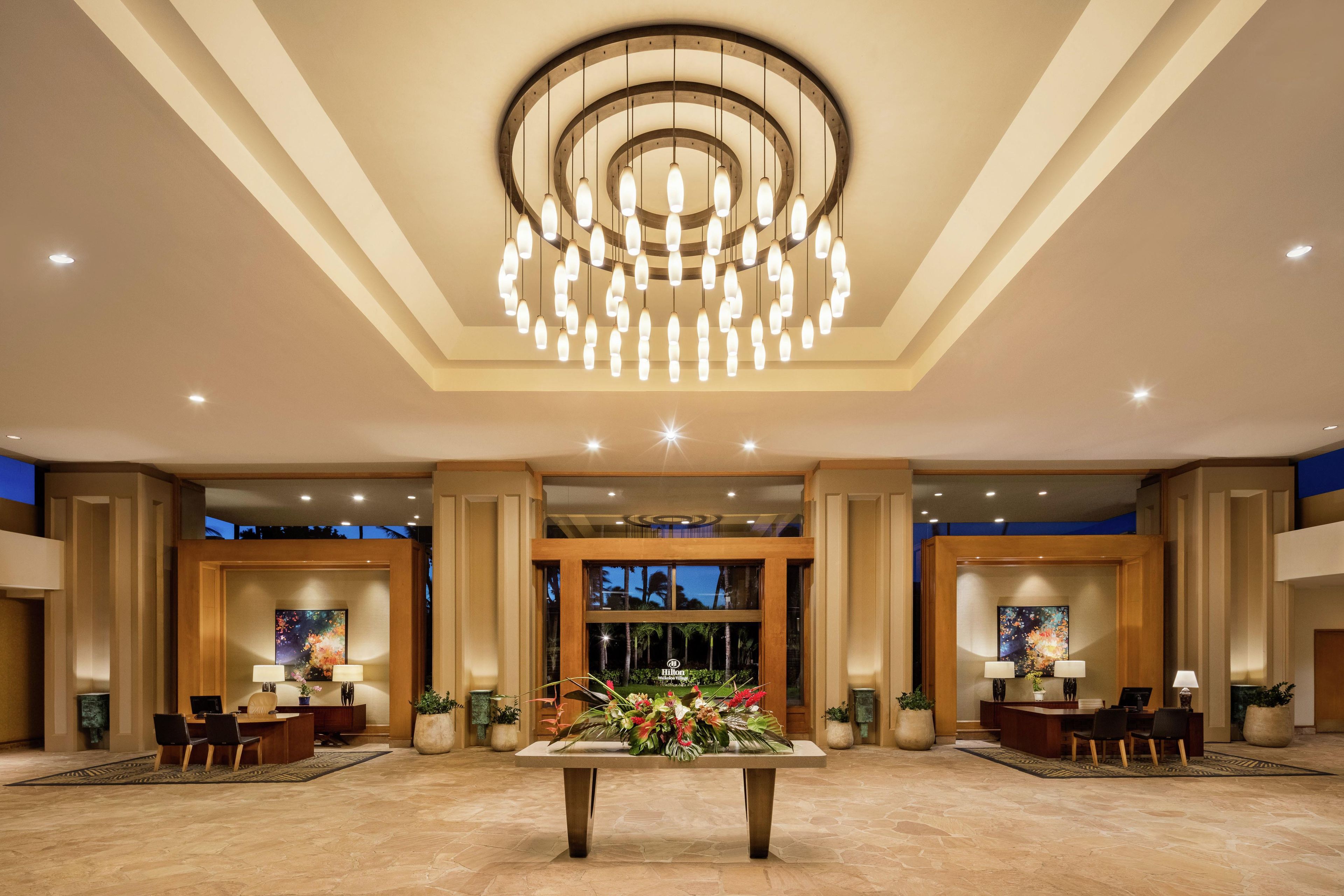A hotel lobby with a large chandelier, two reception desks, and a central floral arrangement.
