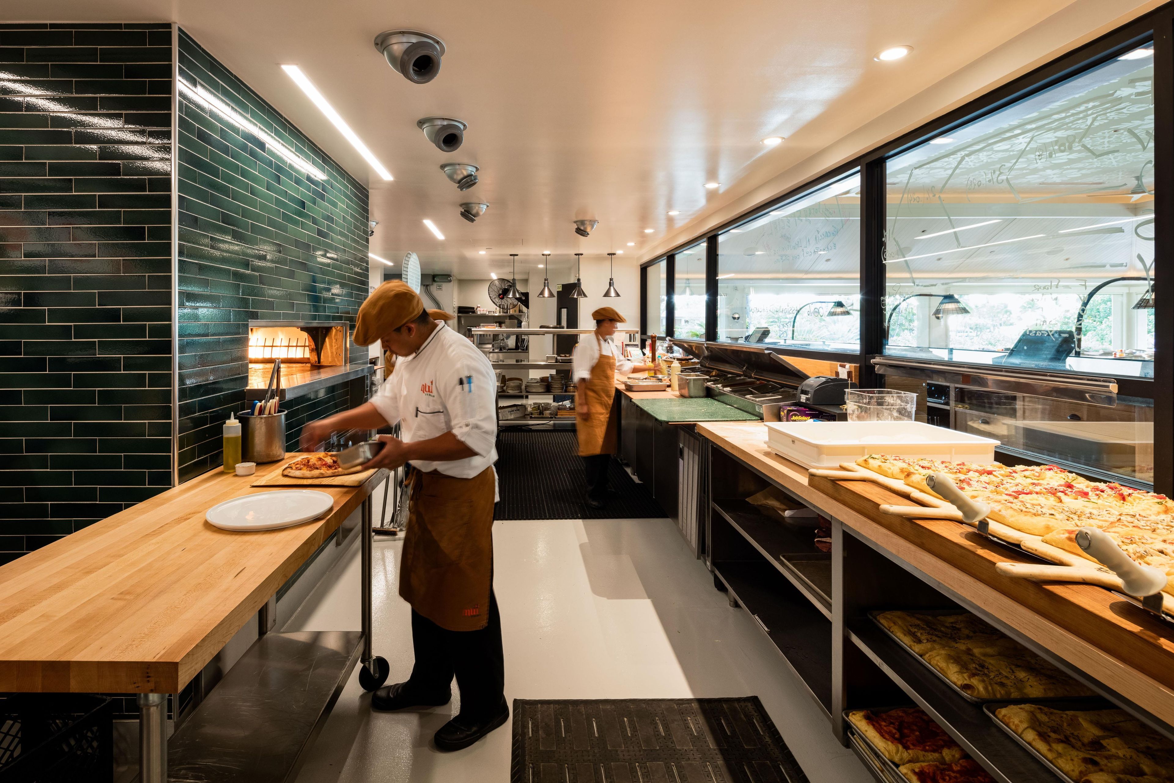 Une cuisine où deux chefs préparent des plats, avec un grand four à pizza et une vue par la fenêtre.