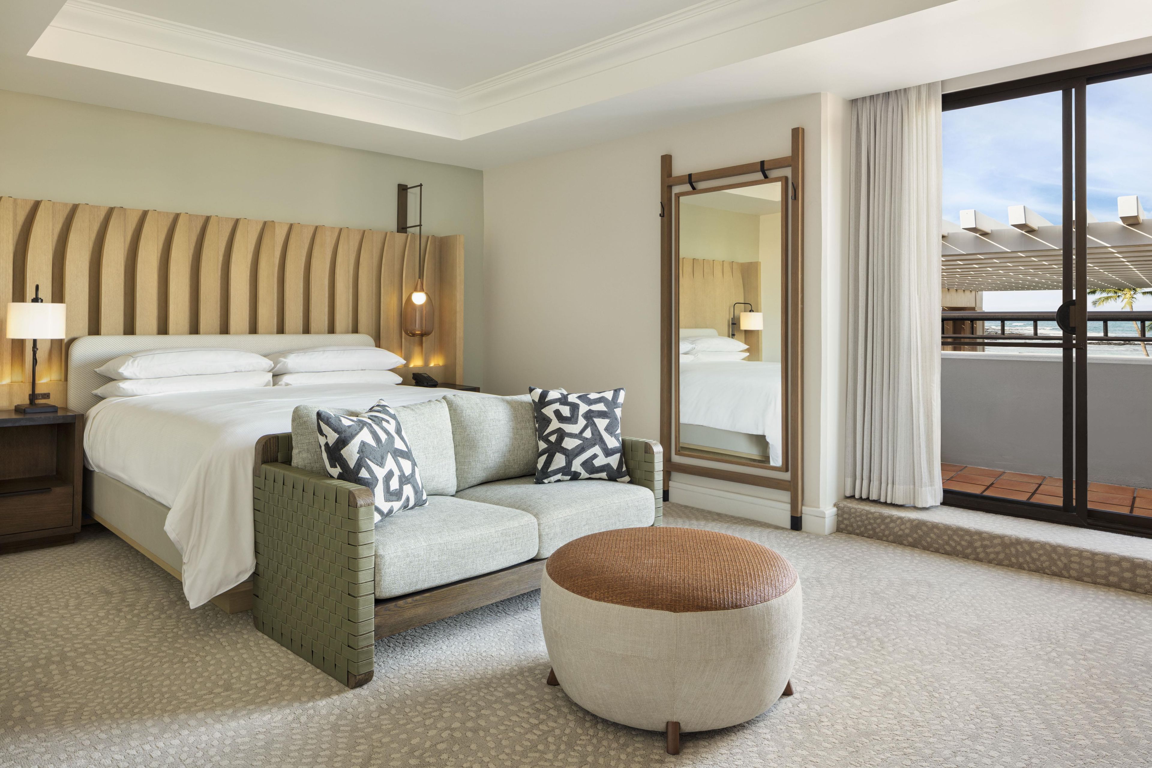 Habitación de hotel con cama, sofá, una otomana redonda, un espejo grande y vista a la playa a través de una ventana.