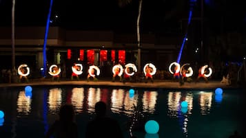 A nighttime poolside with performers using fire hoops, palm trees, and a building with red banners in the background.