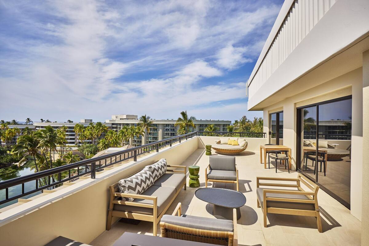 A balcony with a sofa, chairs, a table, and a view of palm trees and buildings.