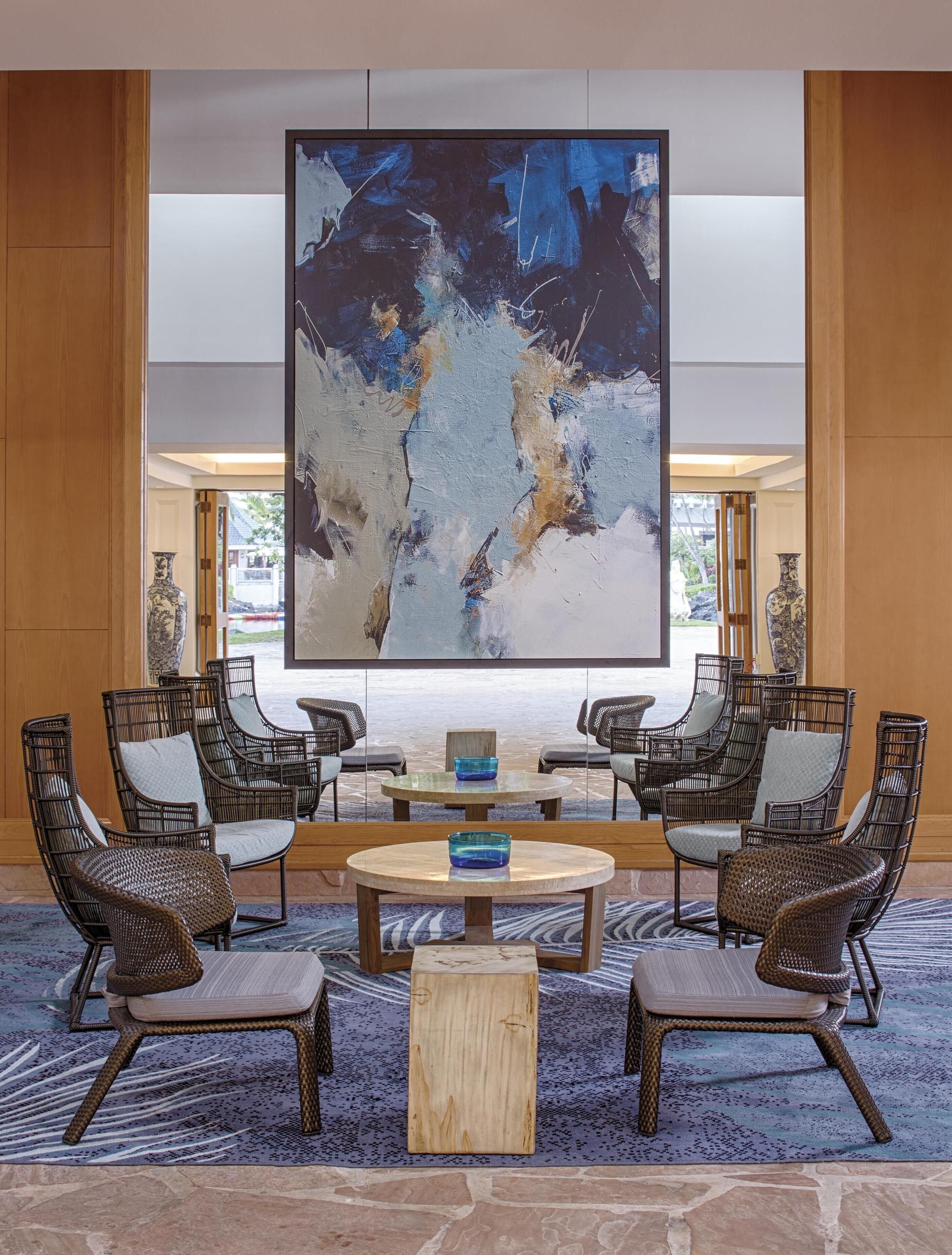 A lobby with a large abstract painting, wicker chairs, a round table, and a stone floor.