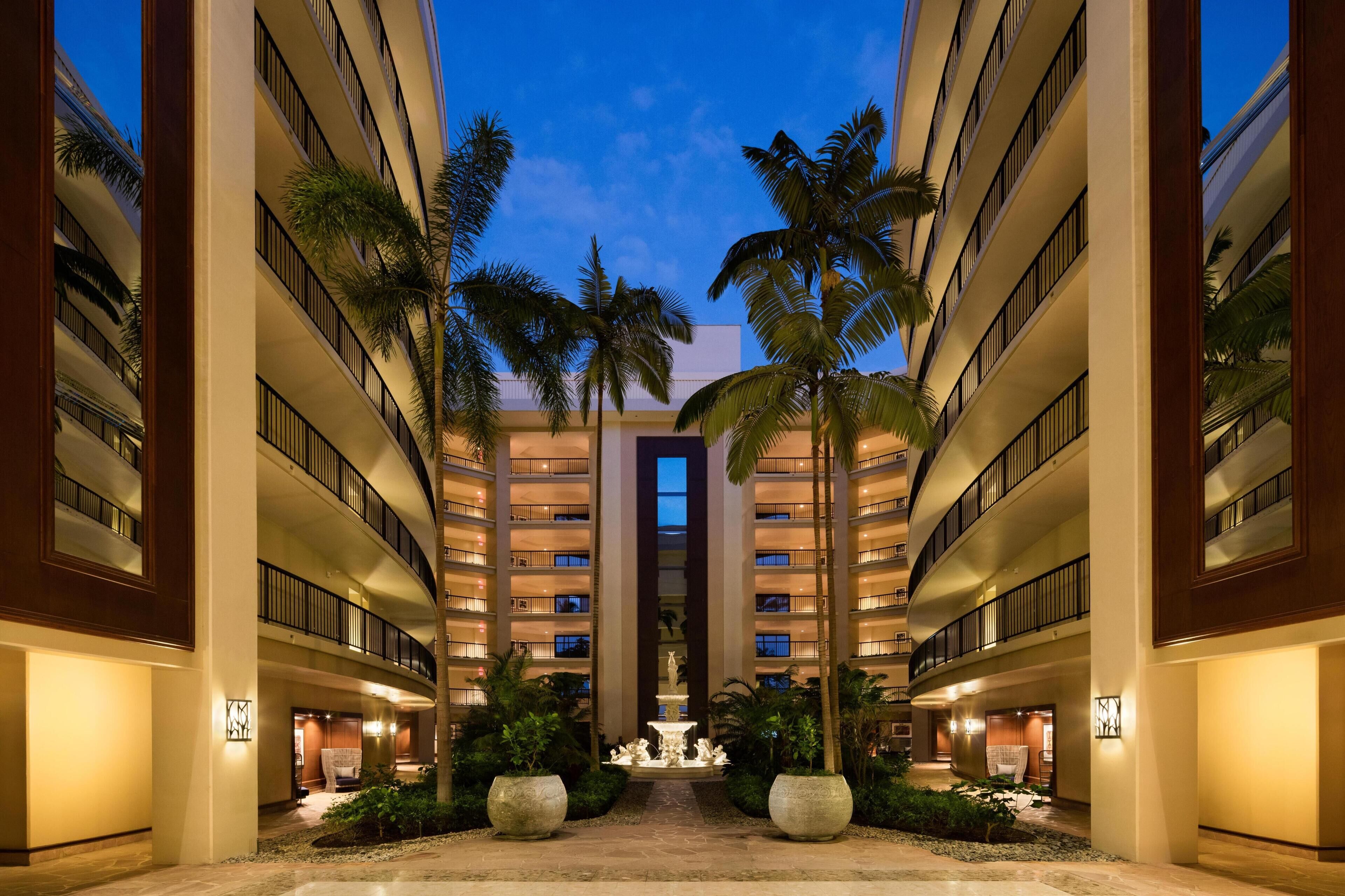 A multi-story hotel with a central fountain, palm trees, and a well-lit courtyard.