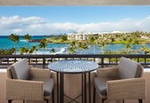 A balcony with wicker chairs and a round table, overlooking a beach with palm trees and a resort building.