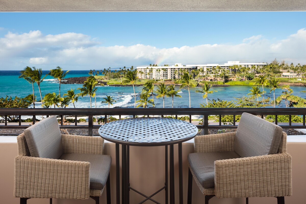 A balcony with wicker chairs and a round table, overlooking a beach with palm trees and a resort building.
