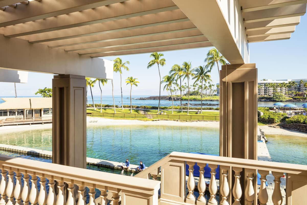 A resort balcony with a view of a pool, palm trees, and a distant beach.