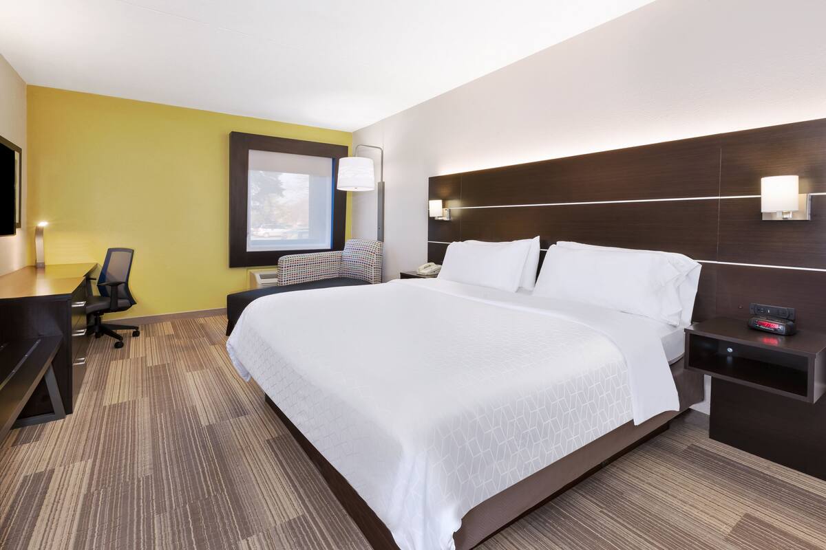A hotel room with a large bed, a desk, a chair, and a nightstand with a lamp.