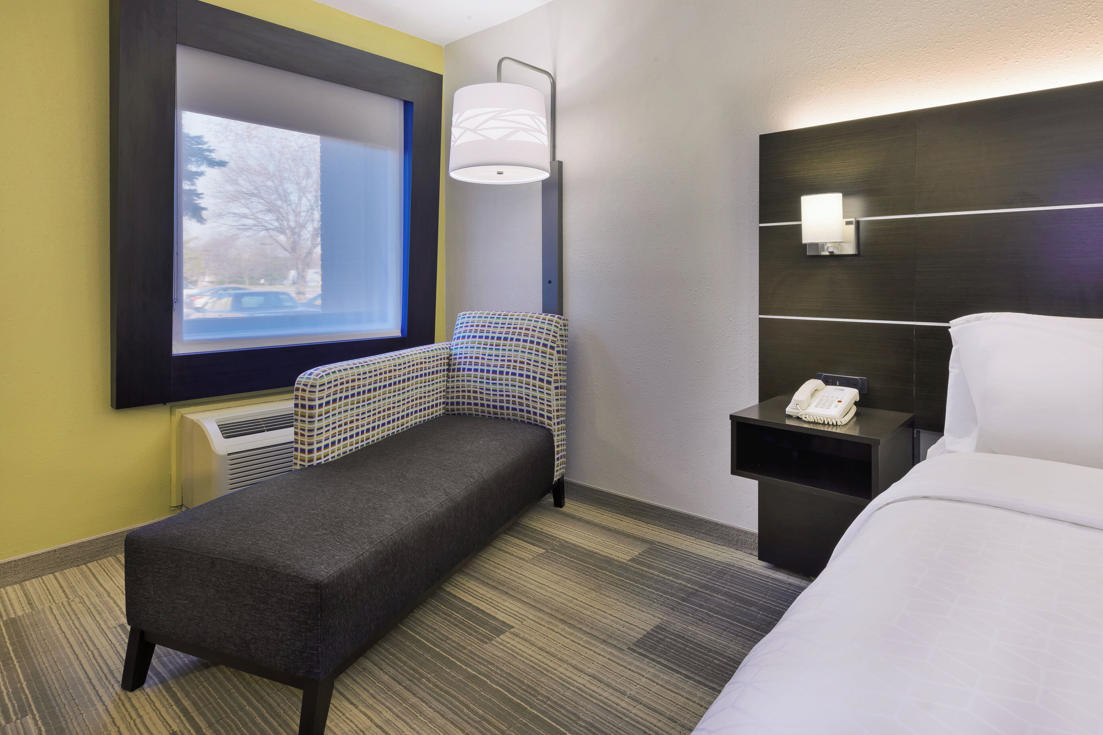 Hotelkamer met een bed, een klein bankje, een nachtkastje met telefoon, een wandlamp en een raam met uitzicht.