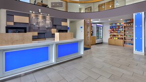 A hotel lobby with a reception desk, a self-service kiosk, a refrigerator stocked with snacks, and a wooden cabinet with various items. - Holiday Inn Express Detroit-Warren by IHG (Warren)