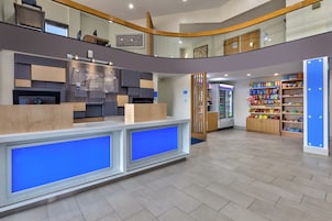 Un vestíbulo de hotel con una mesa de recepción, un quiosco de autoservicio, un refrigerador lleno de refrigerios y un armario de madera con diversos objetos.