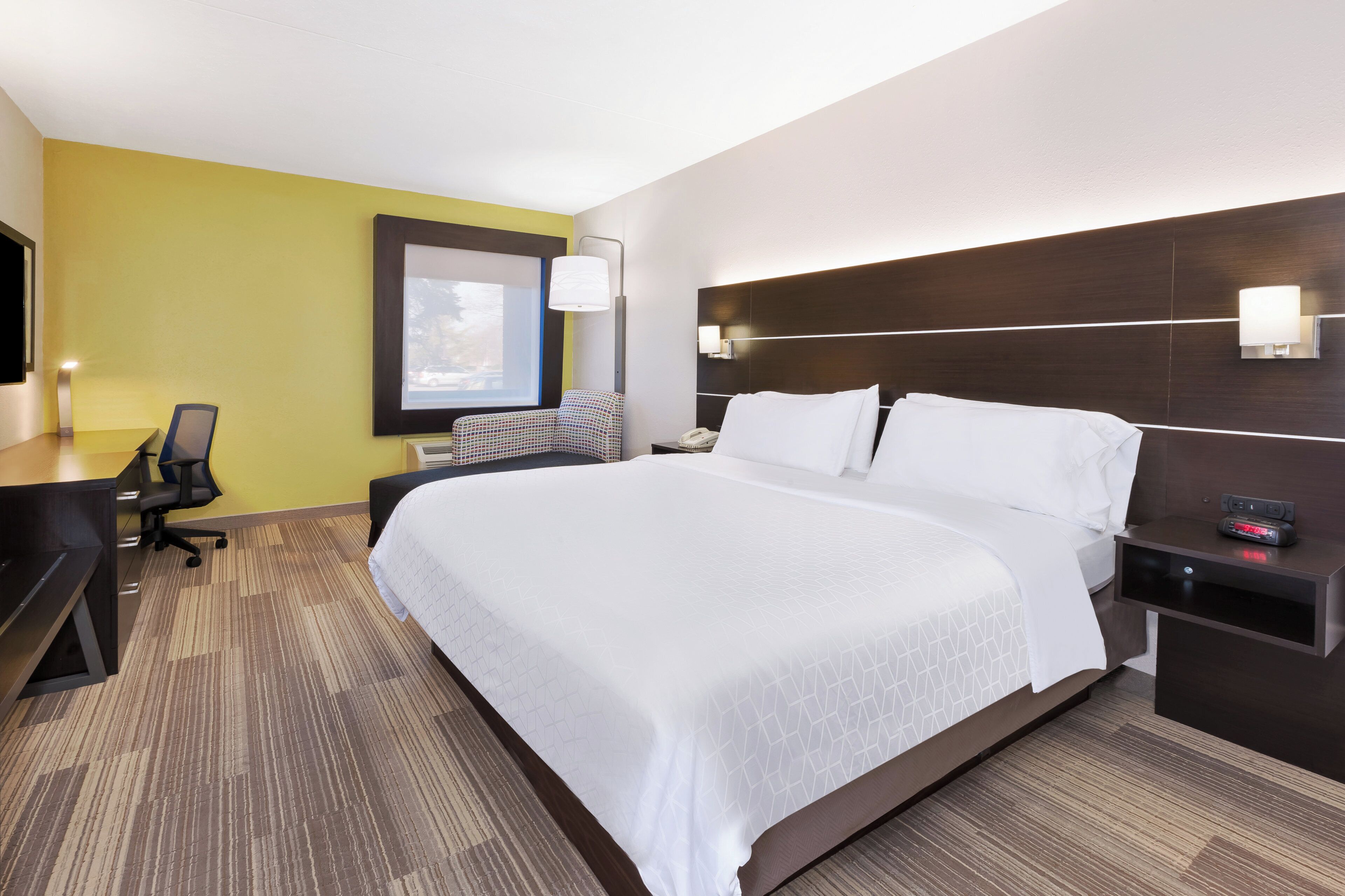 A hotel room with a large bed, a desk, a chair, a nightstand, and a wall-mounted lamp.