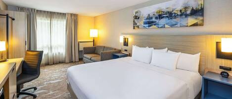 A hotel room with a large bed, a desk with a chair, a small table, and a painting on the wall.
