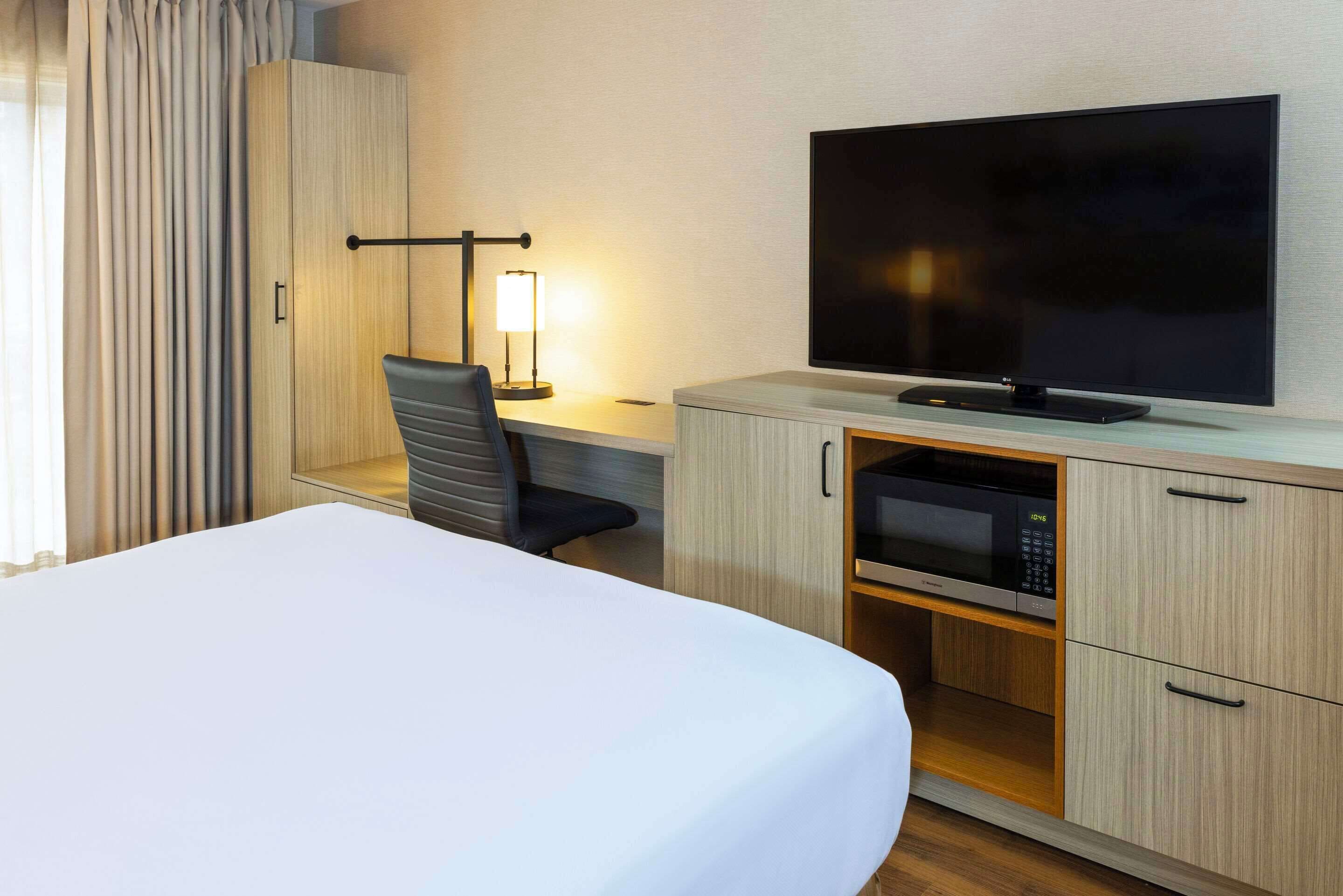 A hotel room with a bed, a desk with a chair, a flat-screen TV, a microwave, and a wardrobe.