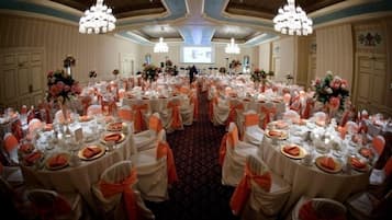 Un salón de banquetes con mesas redondas dispuestas para un evento formal, con manteles blancos y cintas naranjas.
