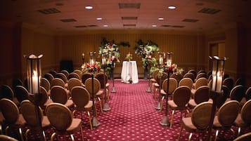 Un salón de conferencias con filas de sillas, un escenario con una mesa y arreglos florales.