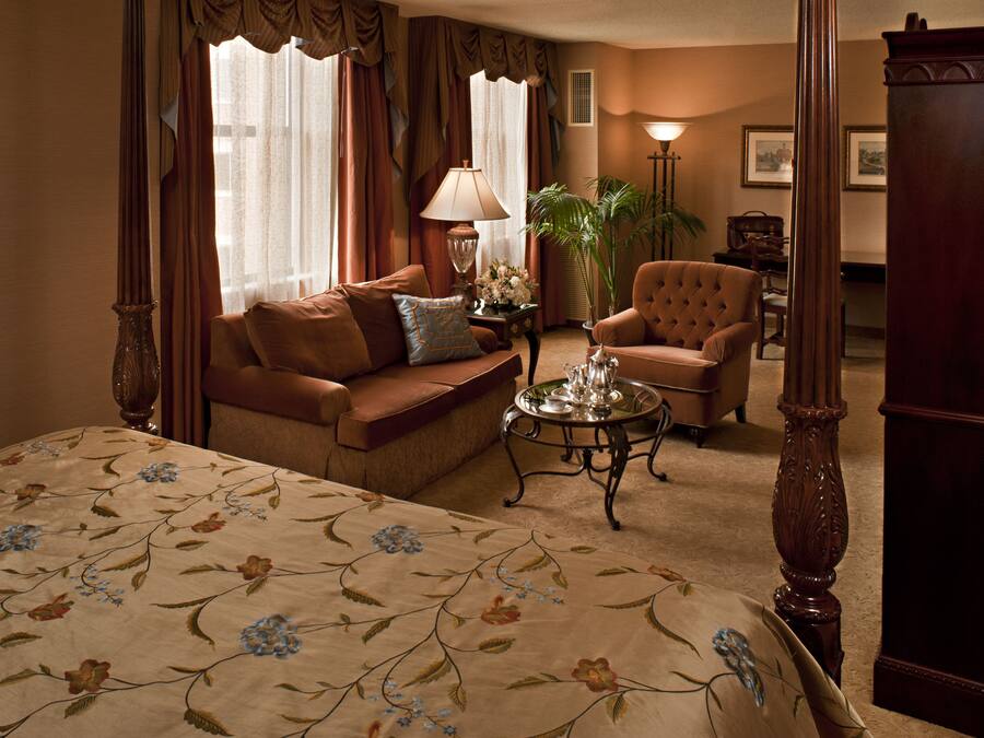 A hotel room with a bed, a sofa, a chair, a table with a tray, curtains, a lamp, and a plant.