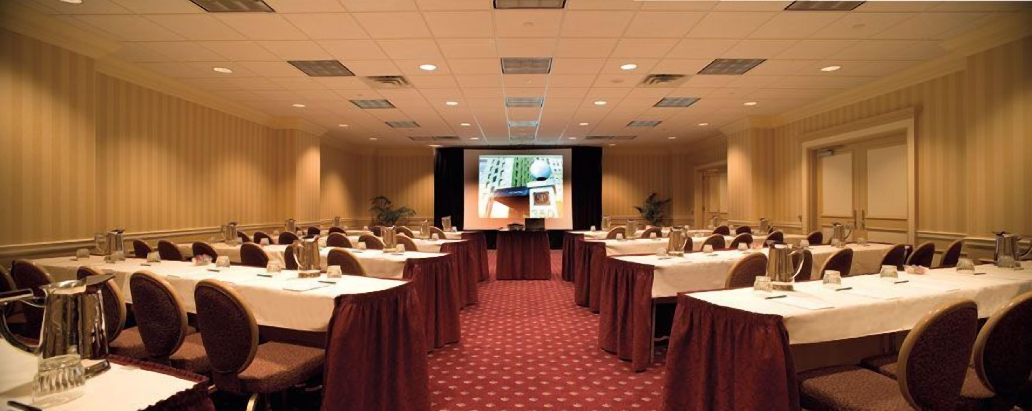Un salón de conferencias con varias mesas, sillas y una pantalla grande al frente.