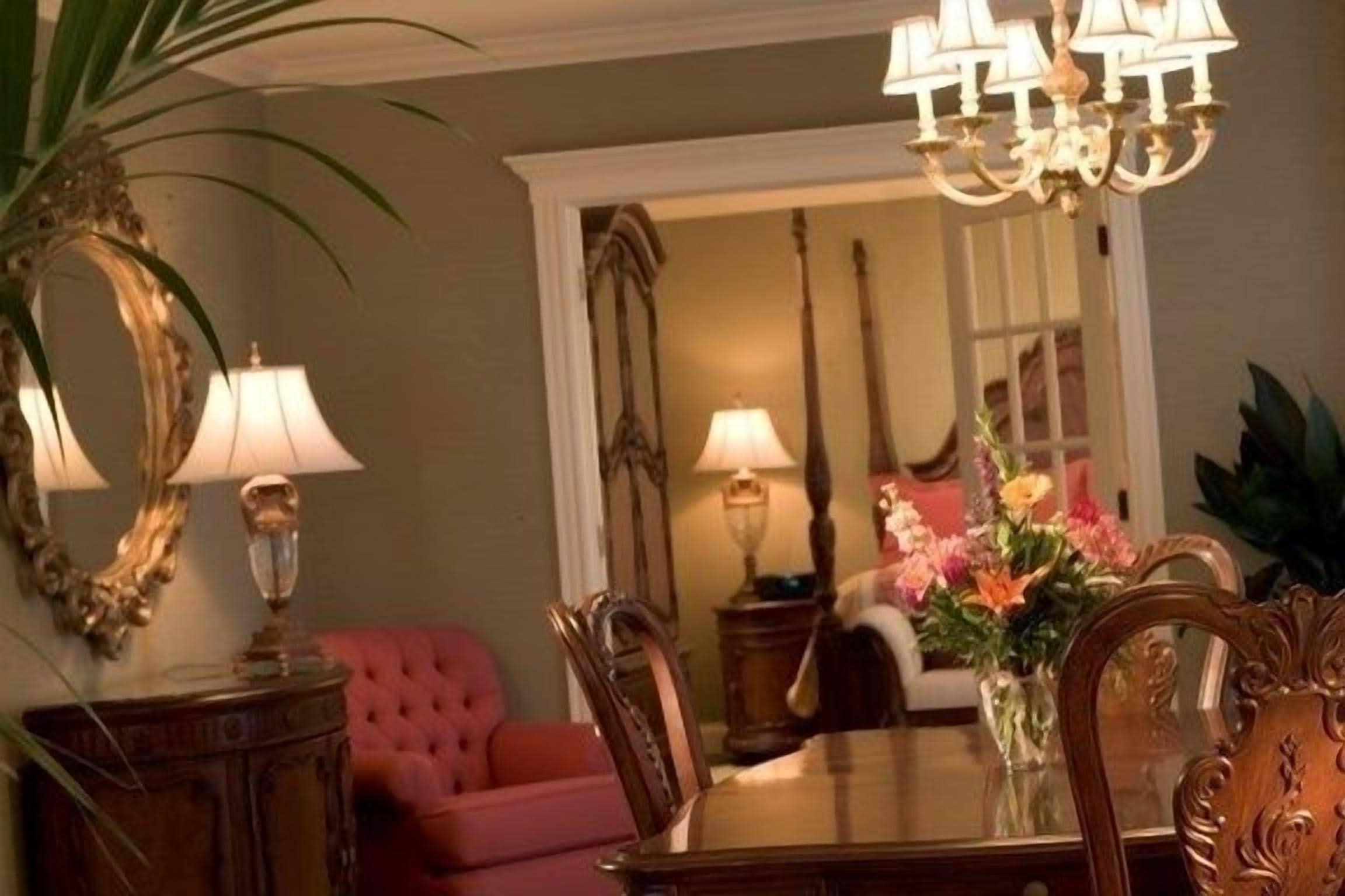 A dining area with a wooden table, chairs, and a chandelier. There is a red sofa and a vase with flowers in the background.
