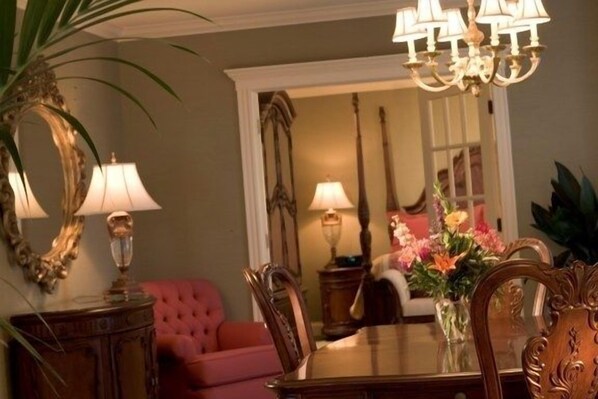 A dining area with a wooden table, chairs, and a chandelier. There is a red sofa and a vase with flowers in the background.