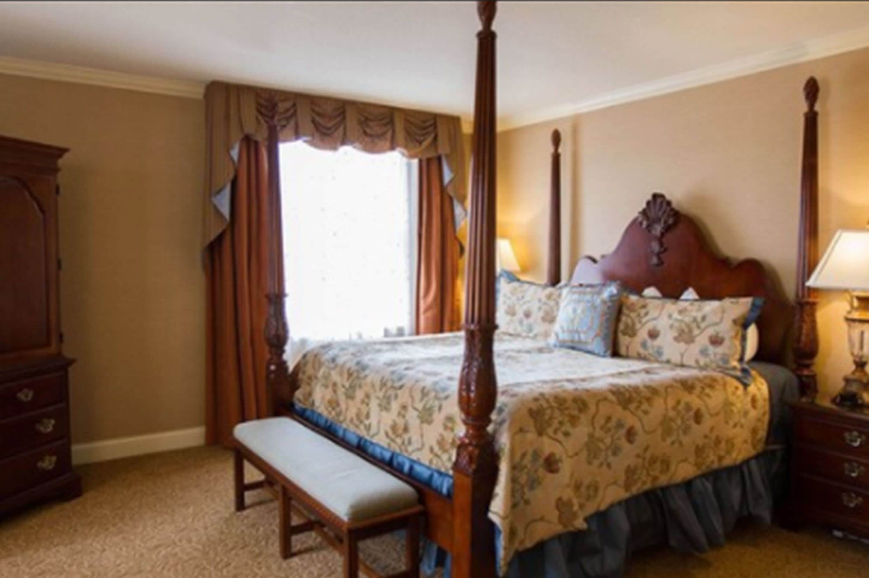 A four-poster bed with a floral bedspread, a wooden bench, and a nightstand with a lamp.