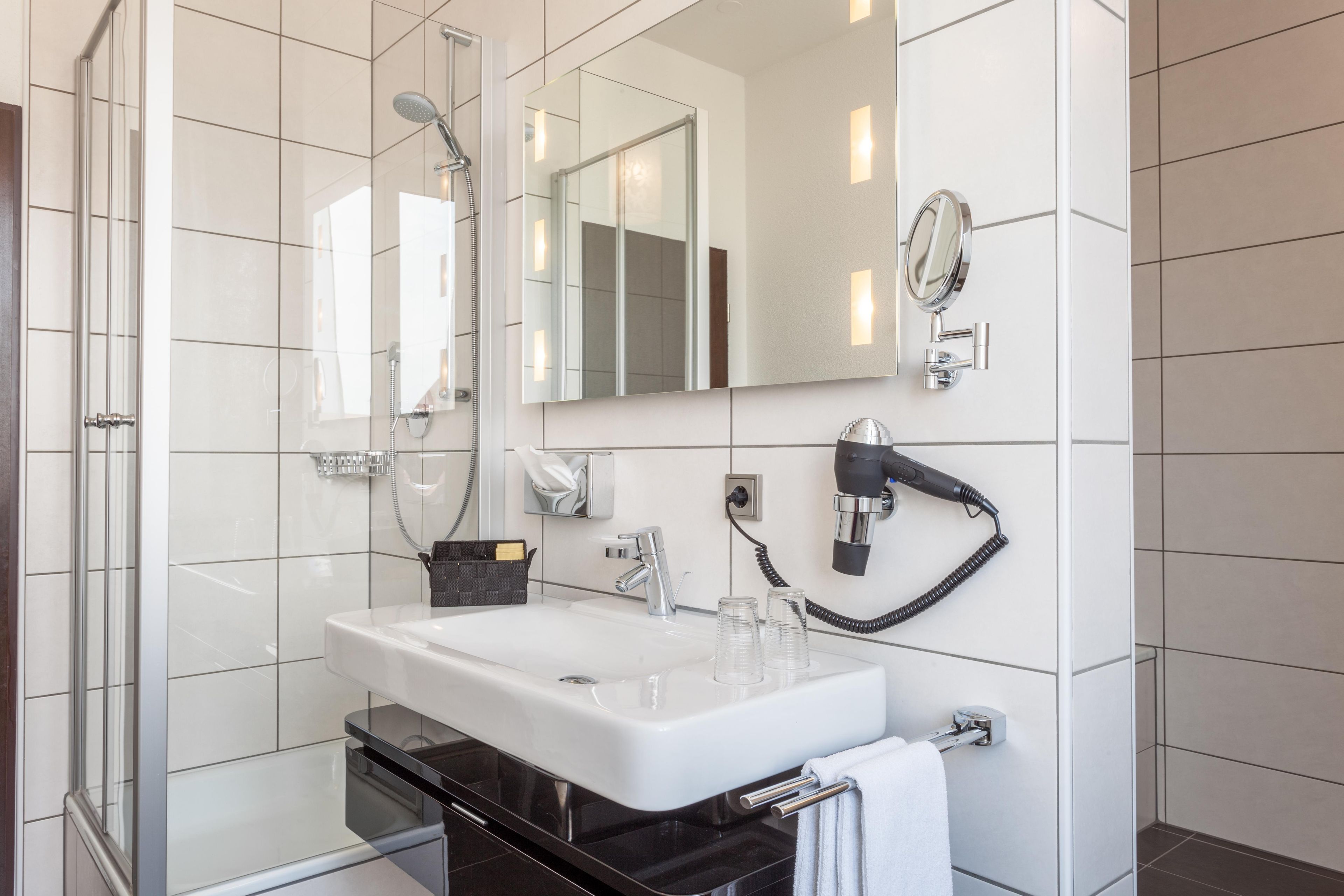 Un baño moderno con cabina de ducha de vidrio, un lavabo blanco y un secador de pelo montado en la pared.