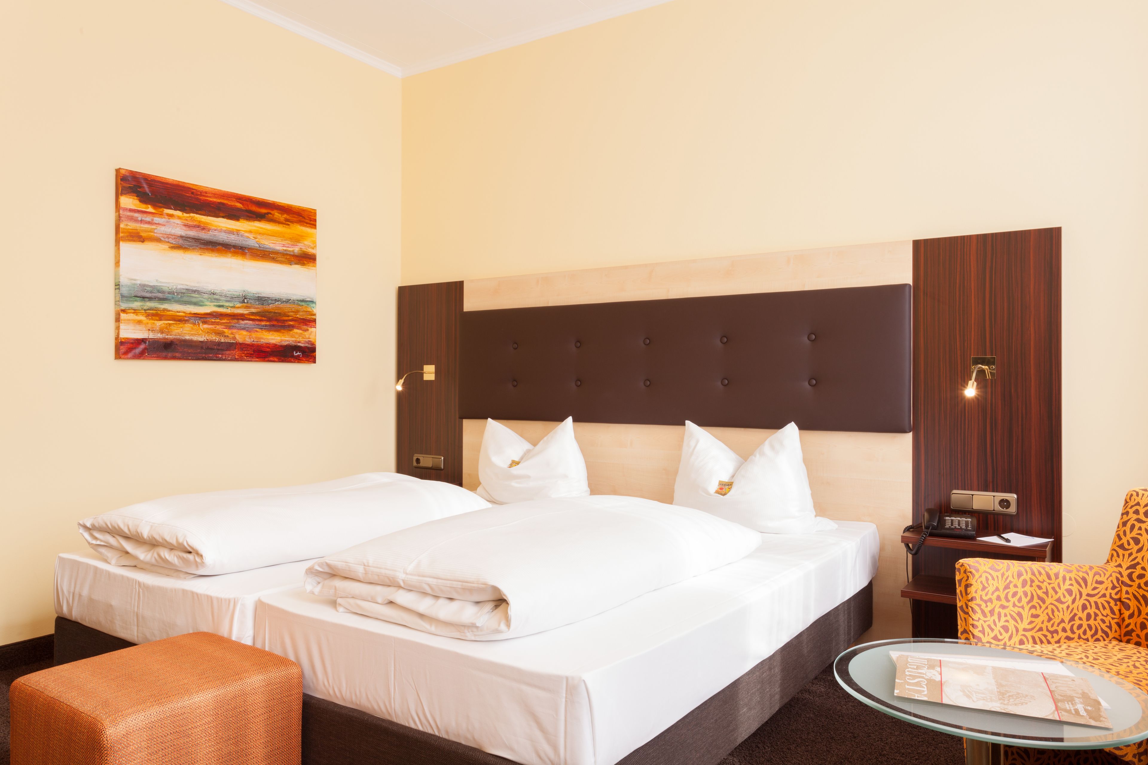 A hotel room with two beds, a wooden headboard, a small table, a chair, and a painting on the wall.