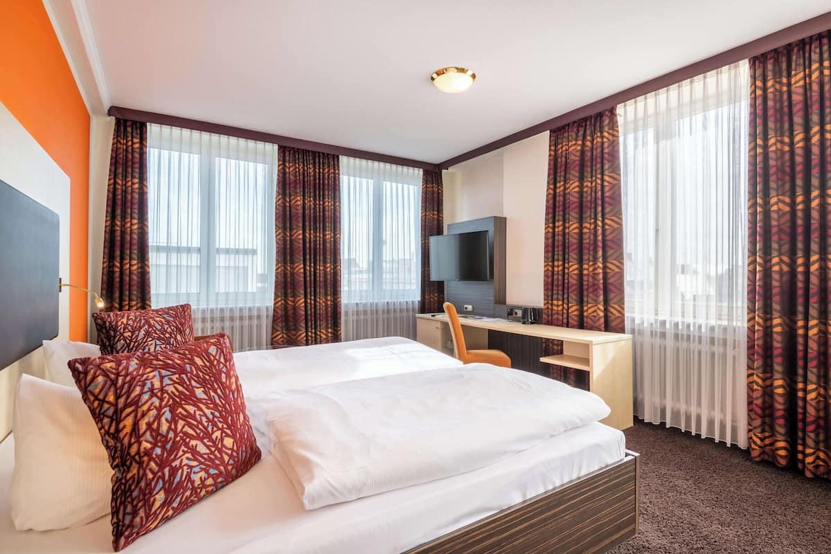 Ein Hotelzimmer mit einem großen Bett, einem Schreibtisch mit Stuhl, einem Fernseher und einem Fenster mit Vorhängen.