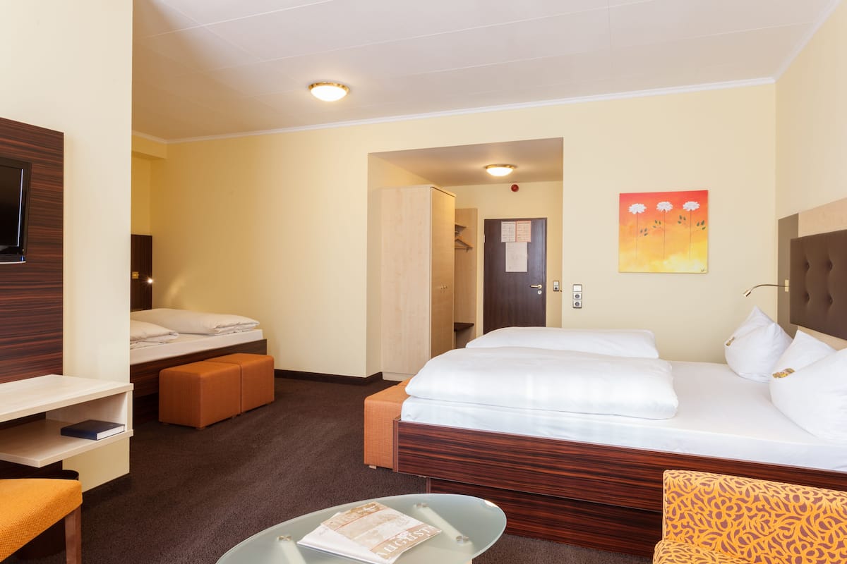 Ein Hotelzimmer mit einem großen Bett, einem Schreibtisch, einem Stuhl und einem kleinen Tisch. Eine Tür führt in einen anderen Raum, und an der Wand hängt ein Bild.