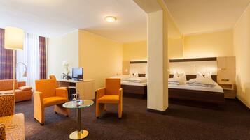 A hotel room with a bed, two armchairs, a small table, a desk with a computer, and a TV.