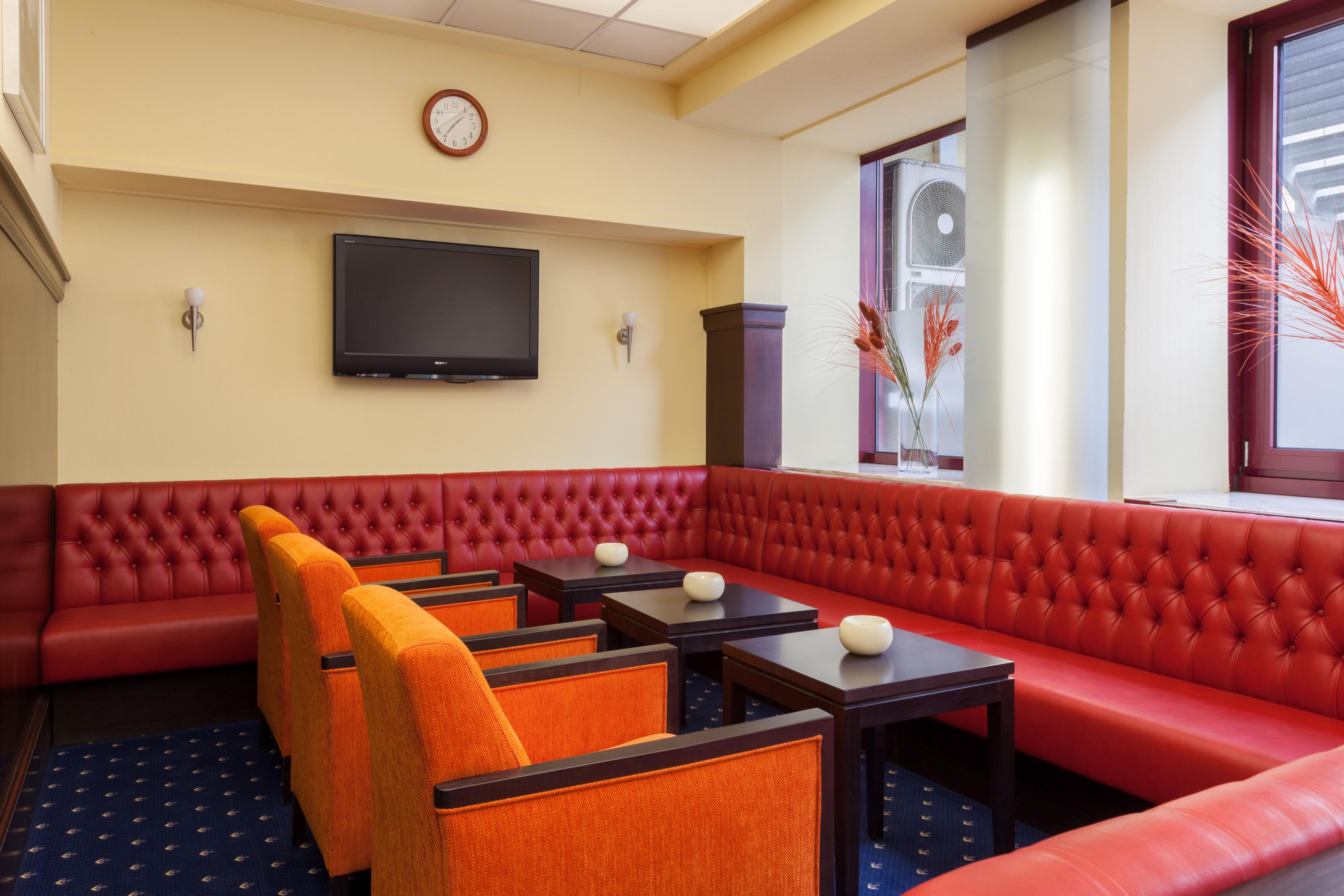 A hotel room with a red upholstered sofa, orange chairs, a flat-screen TV, and a window with decorative plants.