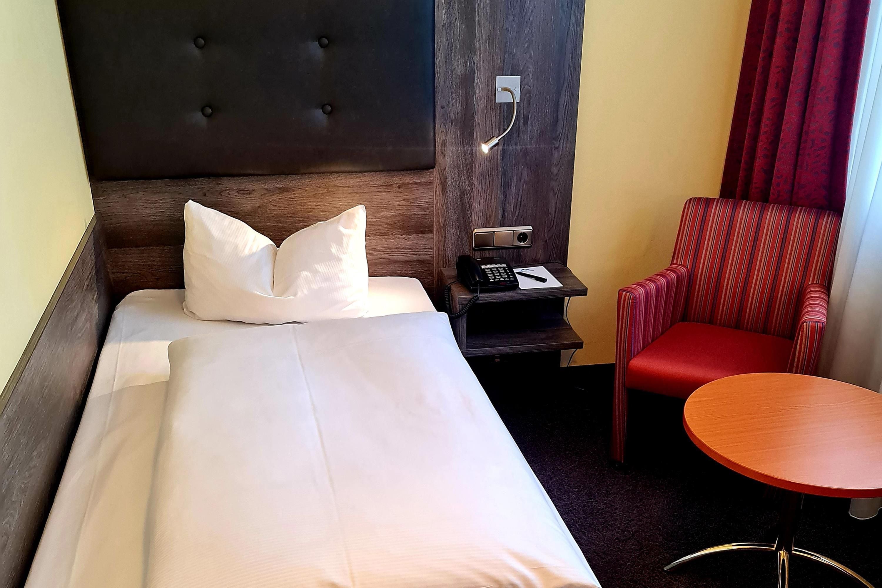 A hotel room with a single bed, a bedside table with a phone and a notepad, a red chair, and a small round table.