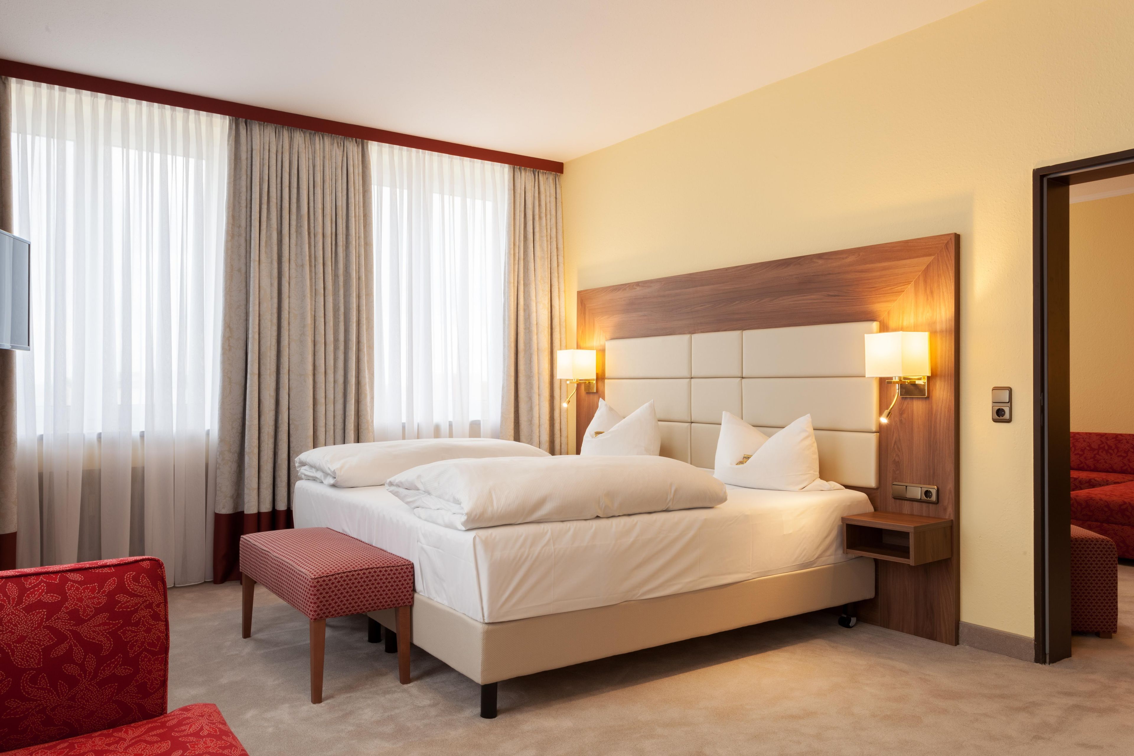 Una habitación de hotel con una cama grande, dos lámparas de noche, un cabecero de madera, una silla roja y una mesita.