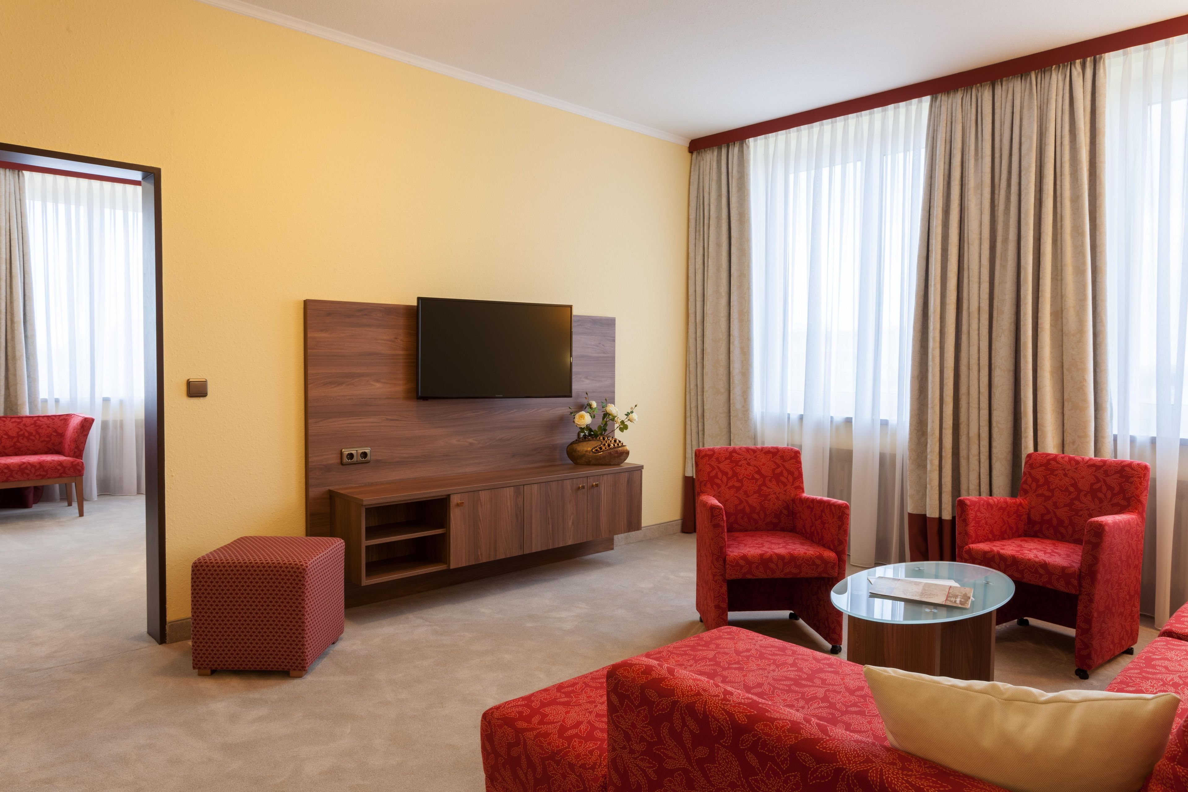 A hotel room with a sofa set, a television, a small table, and a view of the outside through large windows.