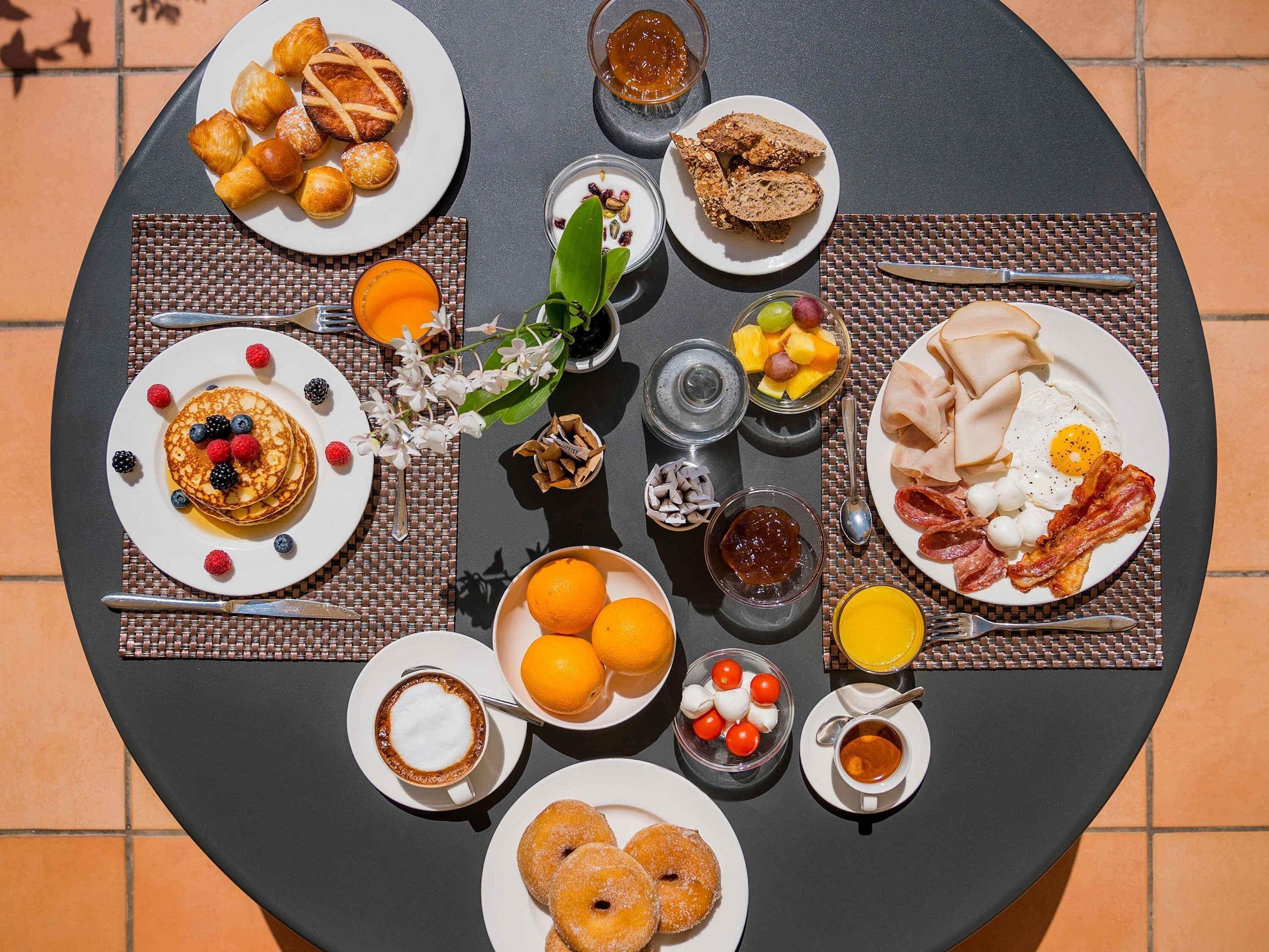 Una tavola per la colazione con diversi piatti, tra cui pancake, croissant, toast, uova, pancetta, salsicce e frutta.