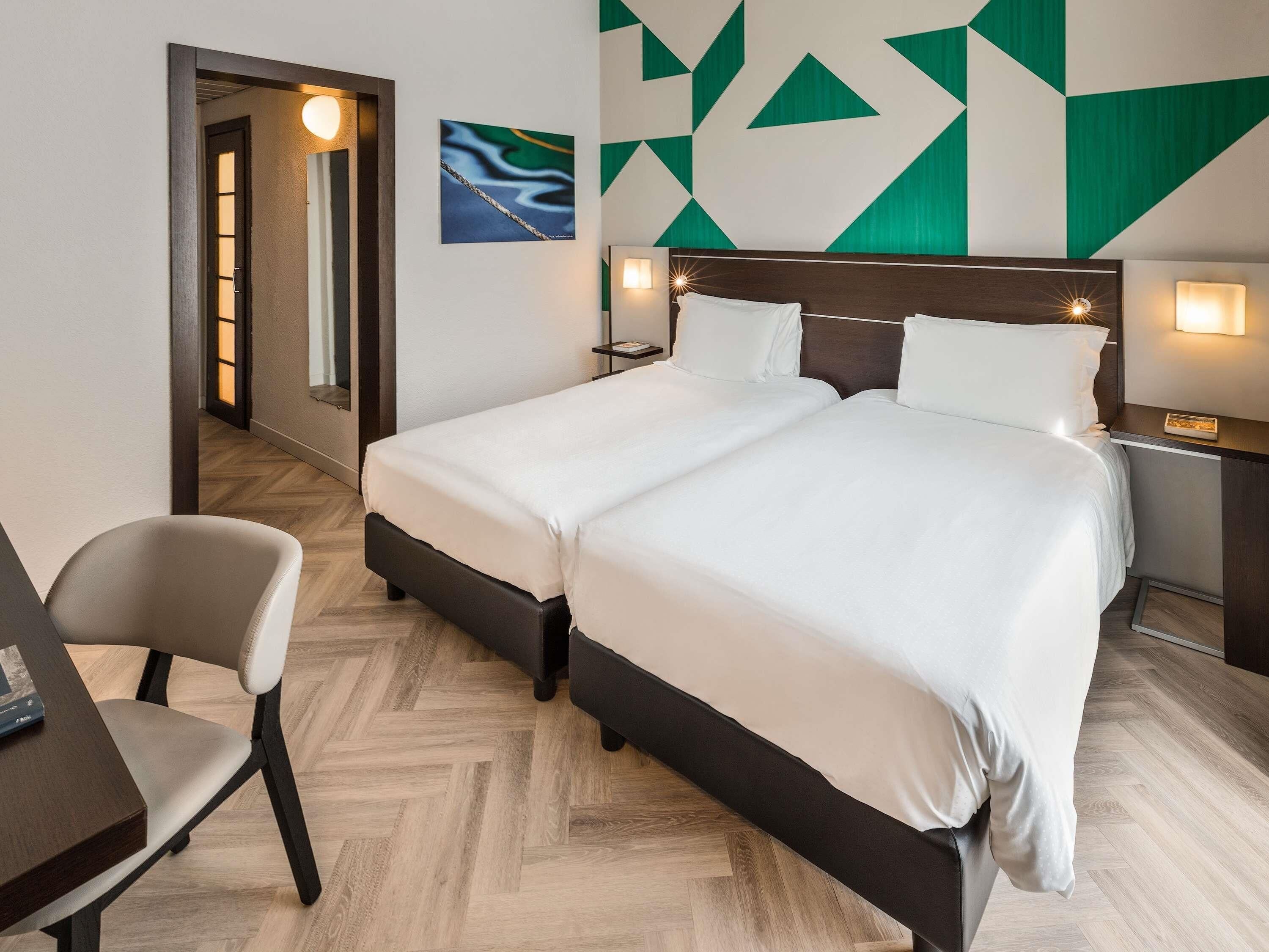 A hotel room with a double bed, two bedside tables with lamps, a desk with a chair, and a herringbone-patterned floor.