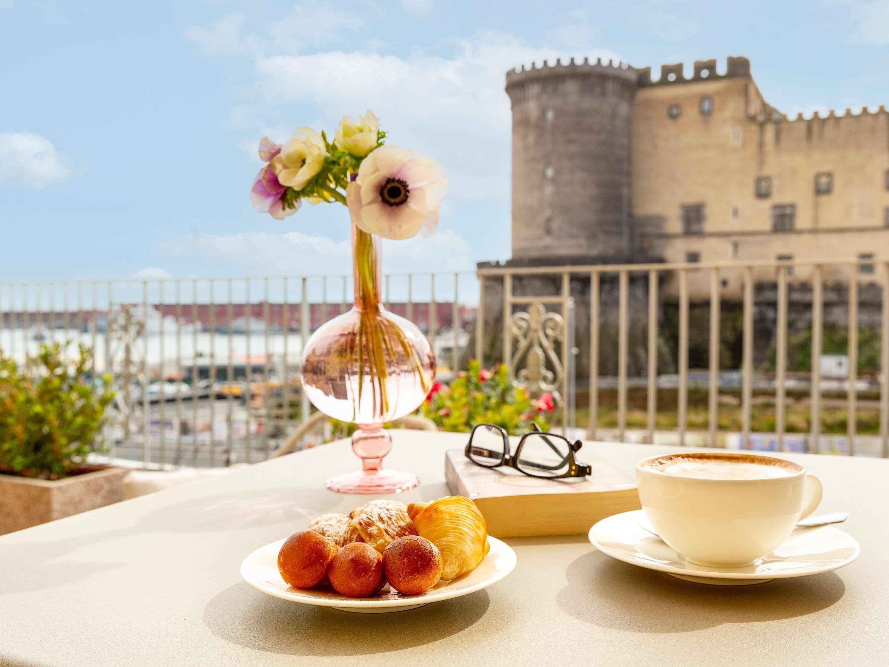 A table with pastries, a cup of coffee, and a vase with flowers, with a castle in the background.
