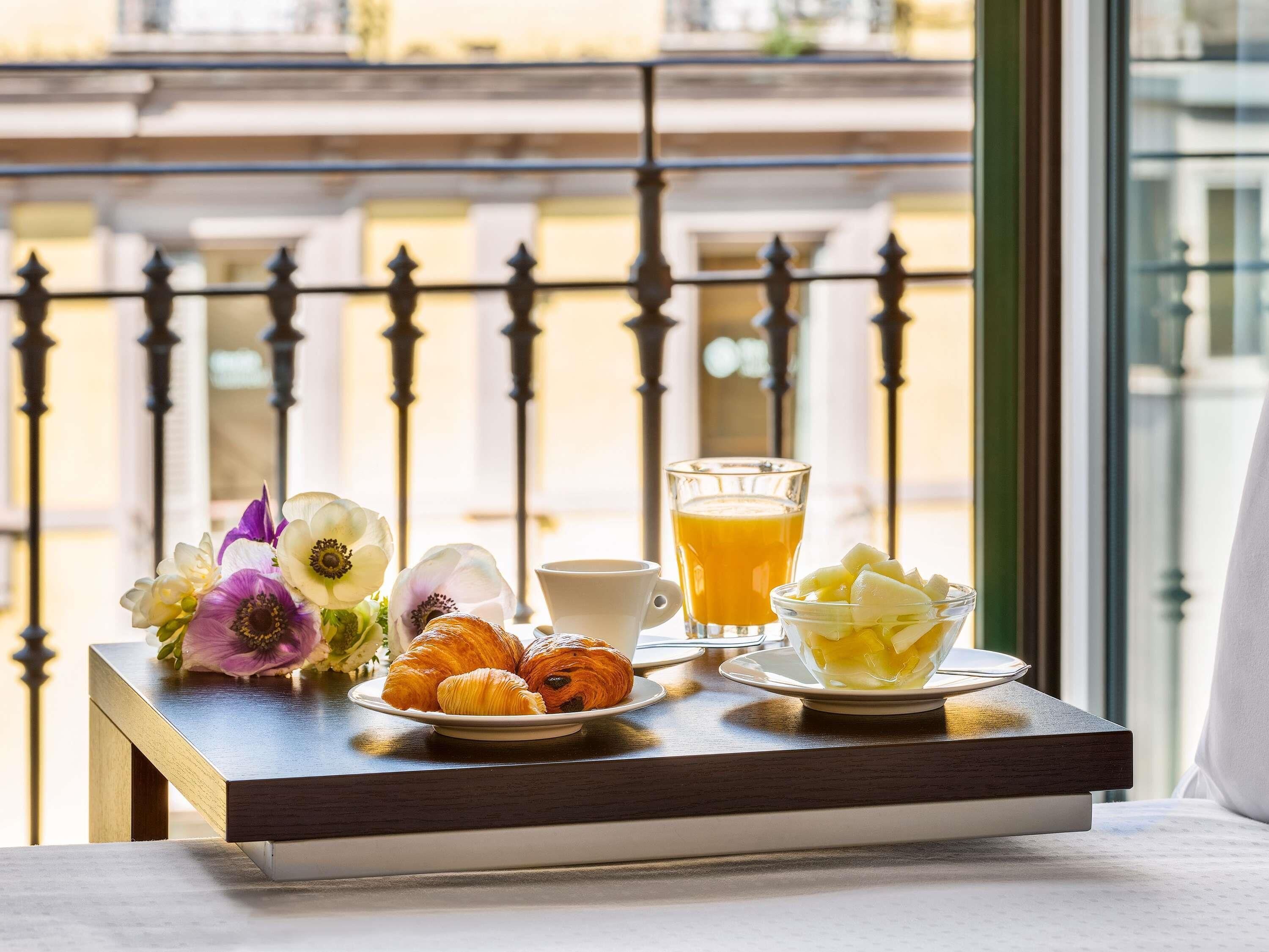 A breakfast setup with croissants, fruit, and orange juice on a table by a window with a balcony view.