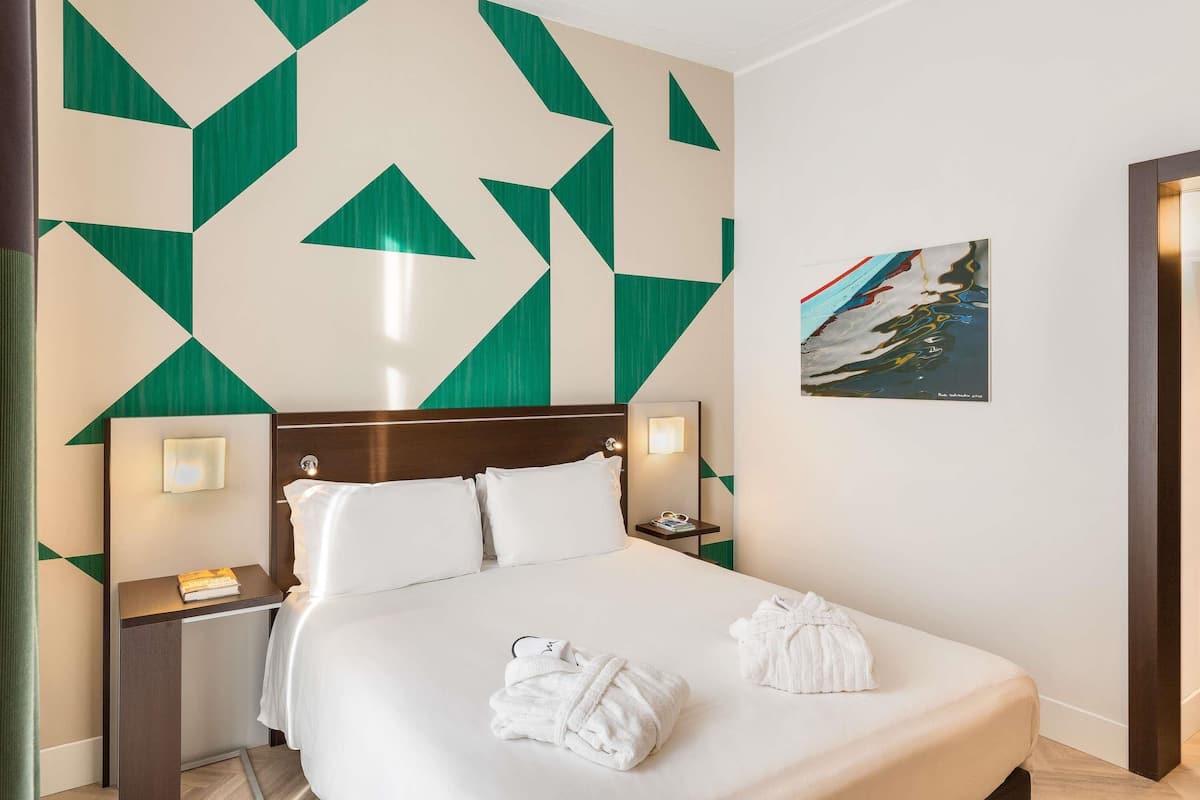 A hotel room with a bed, two bedside tables, a nightstand, a wall with geometric patterns, and a framed picture.