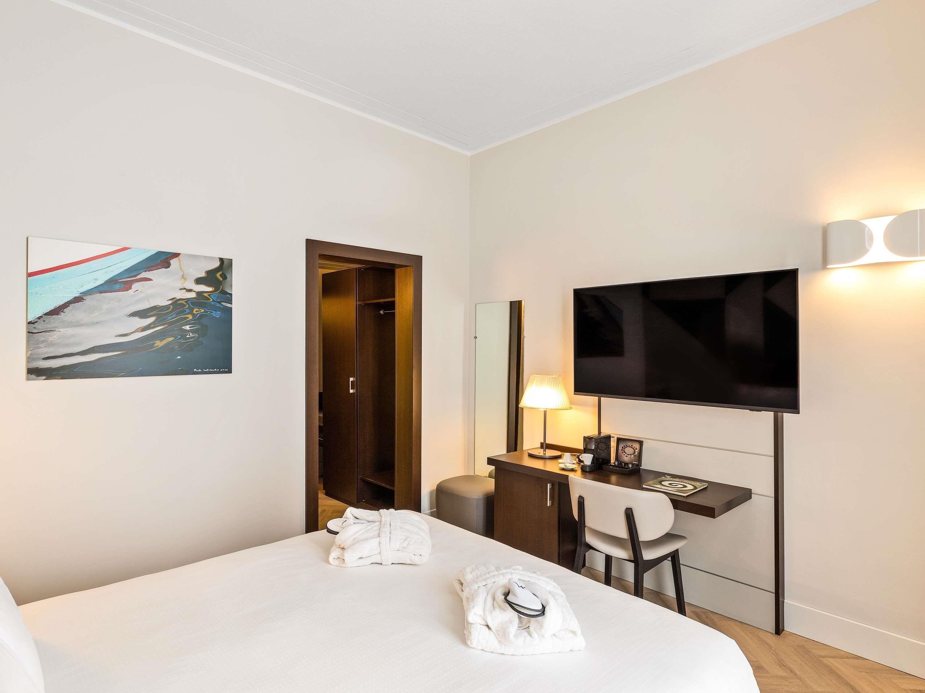 A hotel room with a bed, a desk with a computer, a TV, and a painting on the wall.