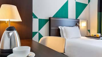 A hotel room with a bed, a bedside table with a lamp, a kettle, and a cup.