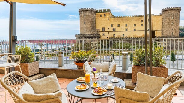Una terraza con sillones de mimbre, una mesa dispuesta con platos de comida y vistas a un castillo.