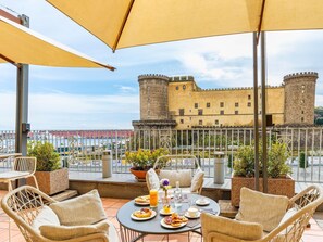 En terrass med flätade stolar, ett bord dukat med mat och utsikt över ett slott.