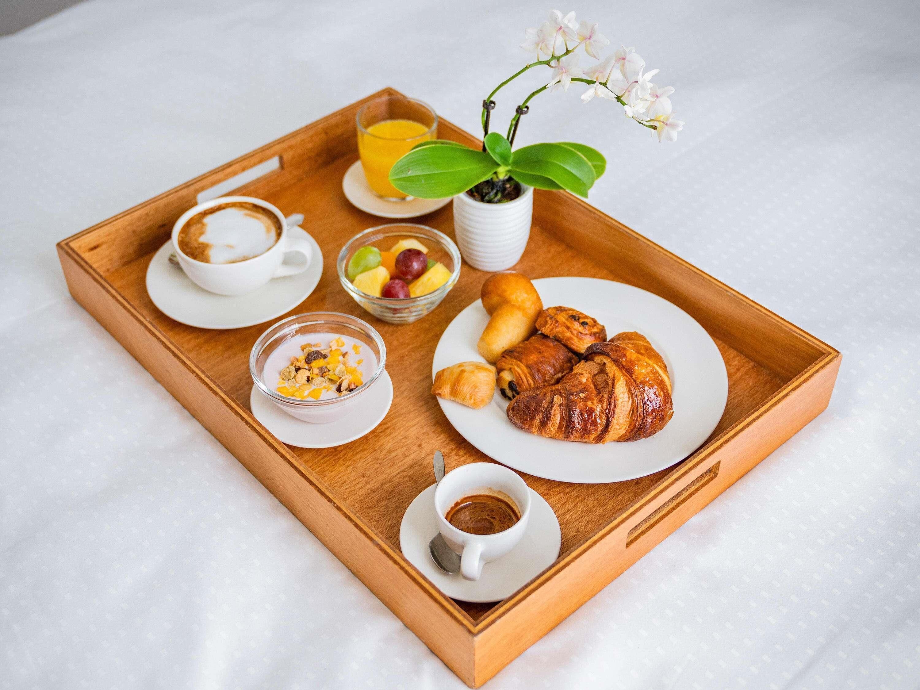 A wooden tray with breakfast items including croissants, fruit, coffee, and a small plant.