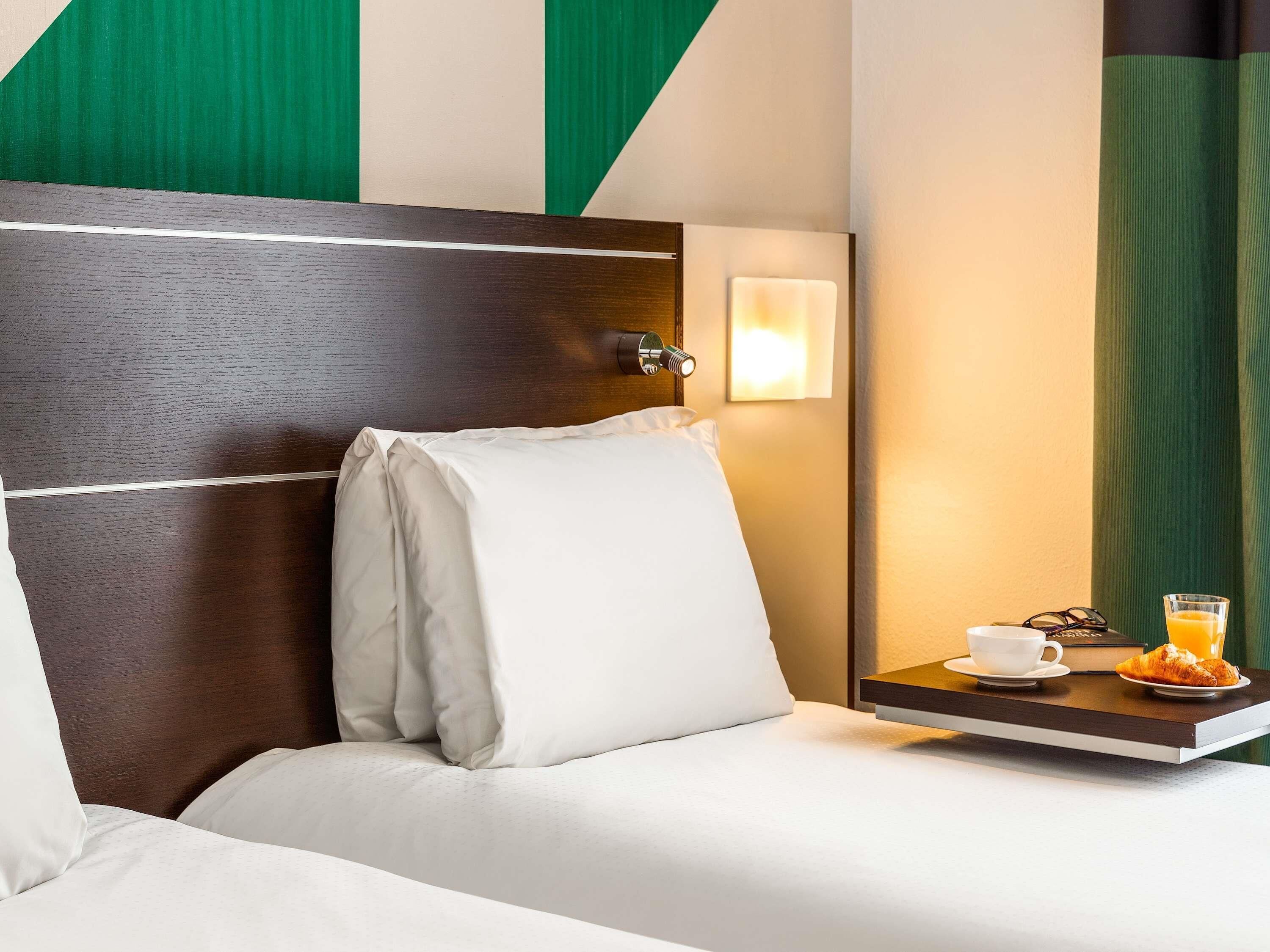 A hotel room with a bed, a bedside table with a cup, a glass of orange juice, and a croissant.